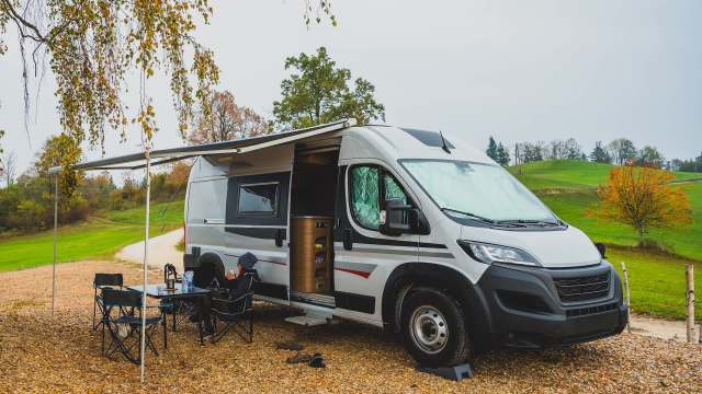 A camper van parked on gravel with a person sitting outside under an awning in a scenic, autumn landscape.