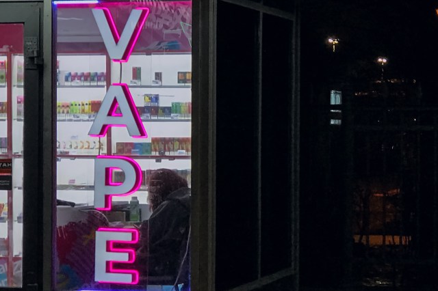 A brightly lit "VAPE" sign in a shop window displays a variety of vape products behind the glass.