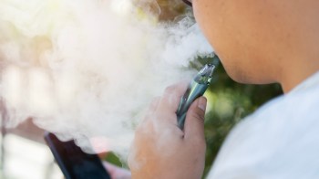 A person vaping outdoors, exhaling vapor while holding a smartphone.