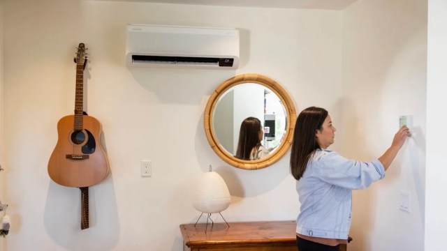 A woman adjusts a thermostat in a room with a guitar, a mirror, and a decorative lamp.