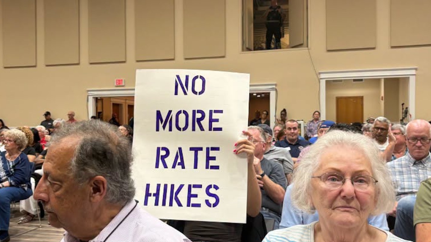 A diverse group of people at a public meeting, with one holding a sign that reads "NO MORE RATE HIKES."