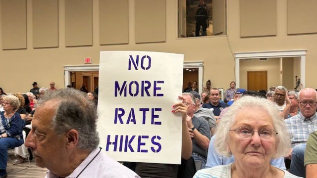 A diverse group of people at a public meeting, with one holding a sign that reads "NO MORE RATE HIKES."