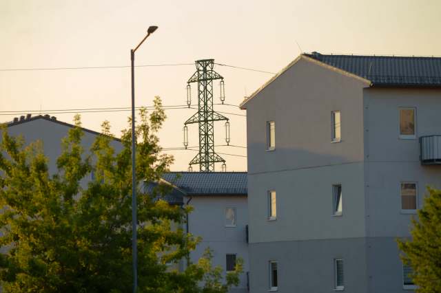 A view of residential buildings with a greenery in the foreground and a power line tower in the background during sunset.
