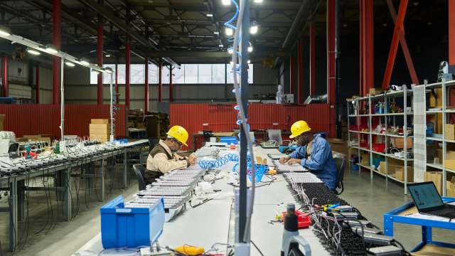 Two workers in yellow hard hats assemble electronic components at a long workbench in a factory setting.