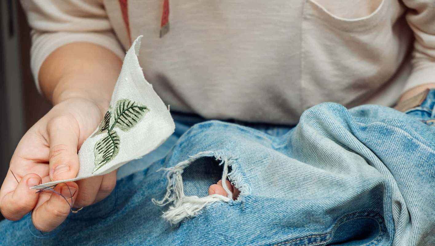 A person holds a piece of fabric with leaf embroidery, sitting next to a ripped pair of jeans.