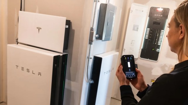 A person using a smartphone app in front of Tesla energy storage units and a circuit panel in a home.
