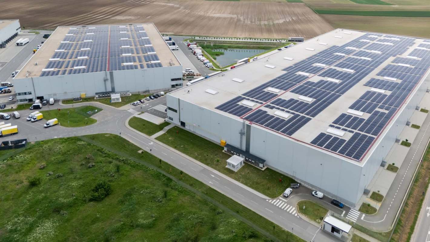 Two large industrial buildings with solar panels on the roofs, surrounded by vehicles and green spaces.