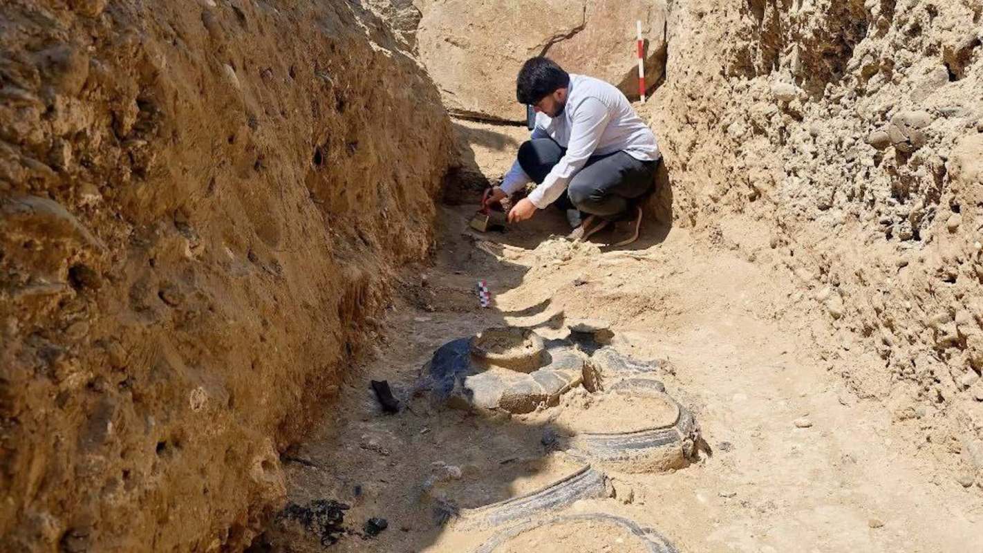 An archaeologist records artifacts while excavating a site with exposed pottery in sandy soil.