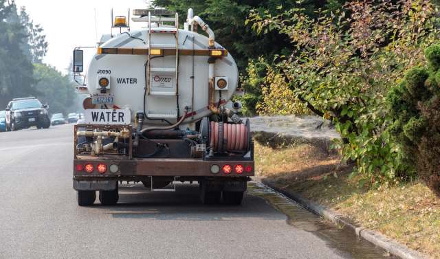 A water truck spraying water along a roadside lined with greenery.
