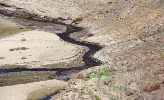 A winding stream through a dry, cracked landscape with sparse vegetation and dry shrubs.
