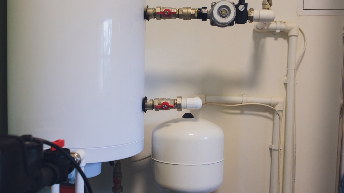 A white water tank connected to pipes and valves on a wall in a mechanical room.