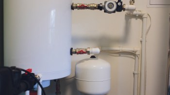 A white water tank connected to pipes and valves on a wall in a mechanical room.