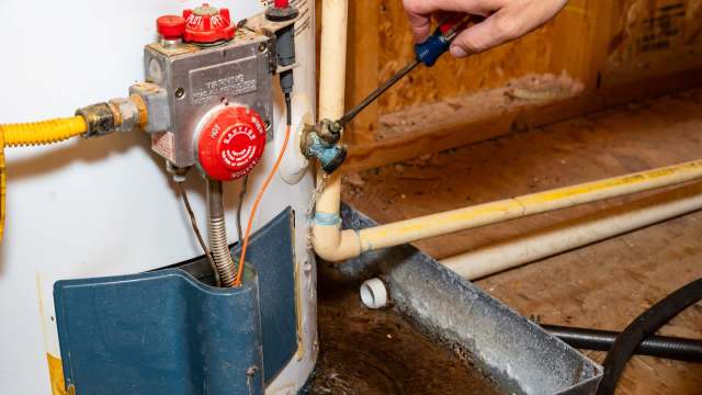 A hand using a screwdriver on a plumbing connection near a leaking water heater with visible water pooling on the floor.
