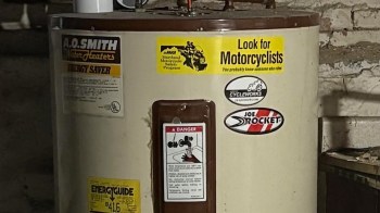 A water heater covered in safety and promotional stickers in a dimly lit basement.