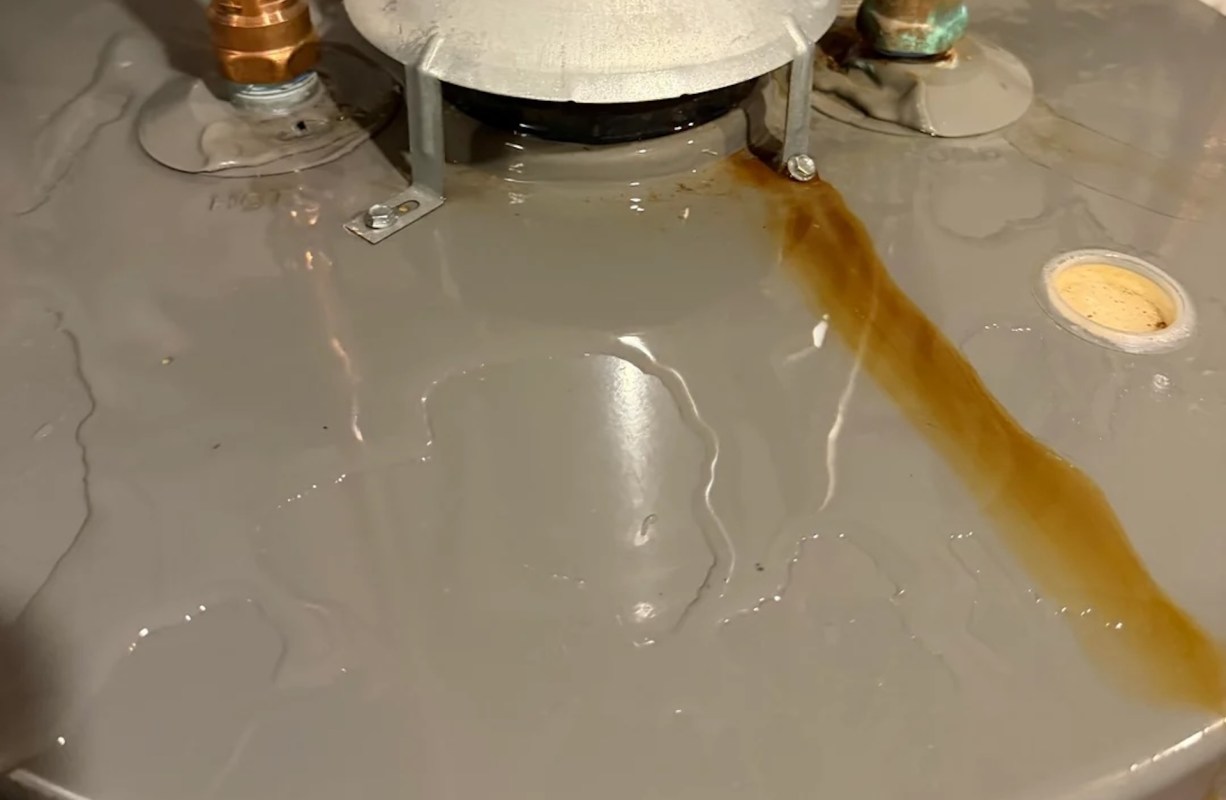 Water is pooling on a gray water heater with a brownish liquid streaking across it.
