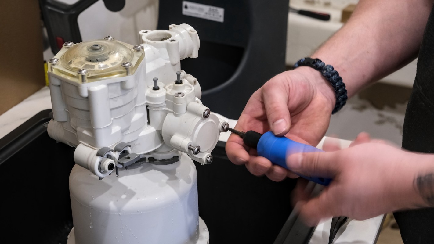 A person uses a blue tool to tighten screws on a white mechanical component in a workshop setting.