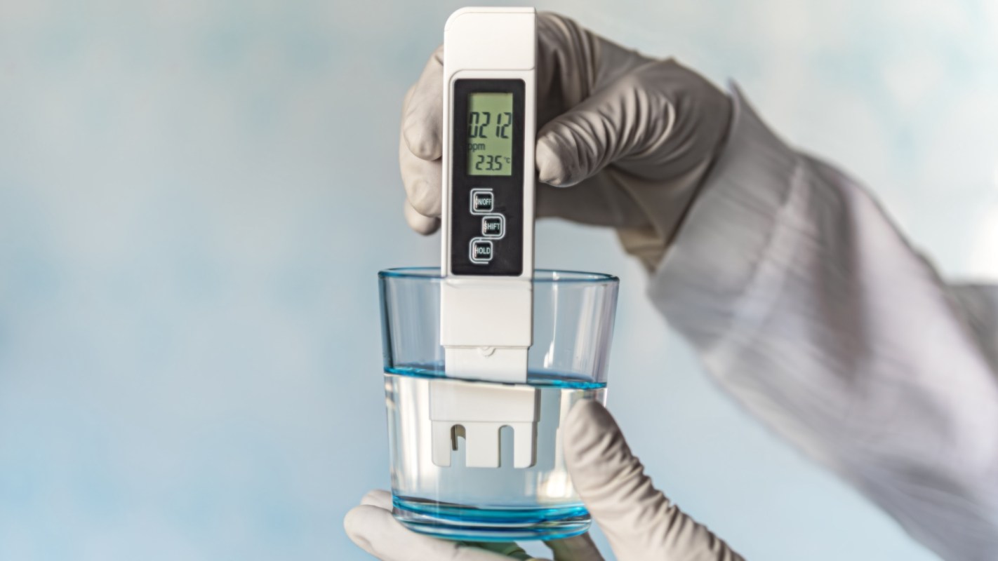 A person in a glove holds a meter above a glass of water, displaying measurements on the screen.