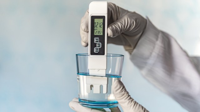A person in a glove holds a meter above a glass of water, displaying measurements on the screen.