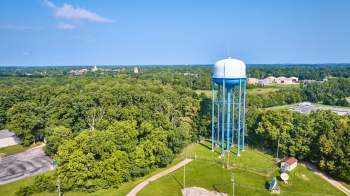 A tall blue water tower surrounded by dense greenery and a distant view of buildings under a clear blue sky.