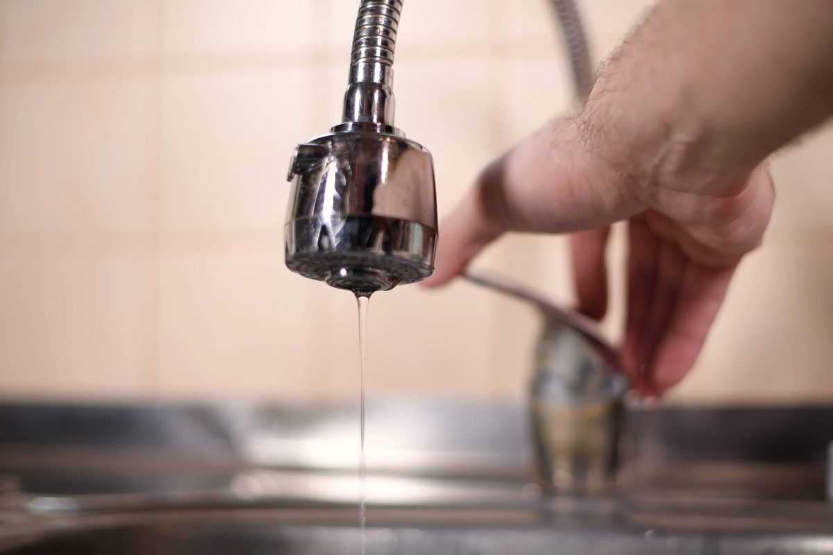 A close-up of a faucet dripping water with a hand adjusting another faucet in the background.