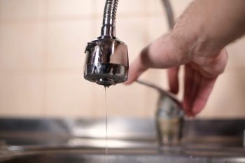 A close-up of a faucet dripping water with a hand adjusting another faucet in the background.