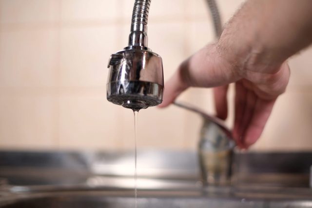 A close-up of a faucet dripping water with a hand adjusting another faucet in the background.