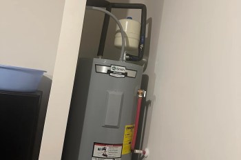 A gray water heater with labeled hoses and a nearby blue basin in a small, enclosed space.