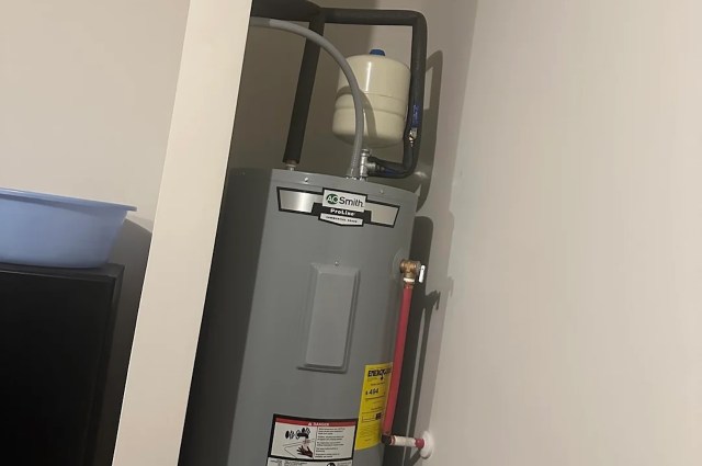 A gray water heater with labeled hoses and a nearby blue basin in a small, enclosed space.