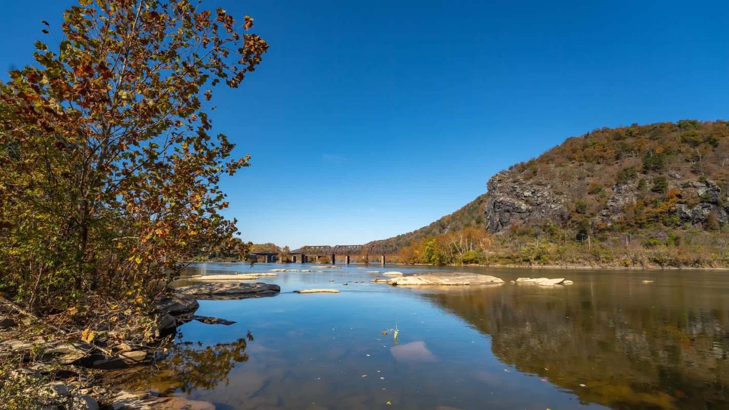 A river landscape with rocky shores, a bridge in the background, and colorful autumn foliage on the hills.