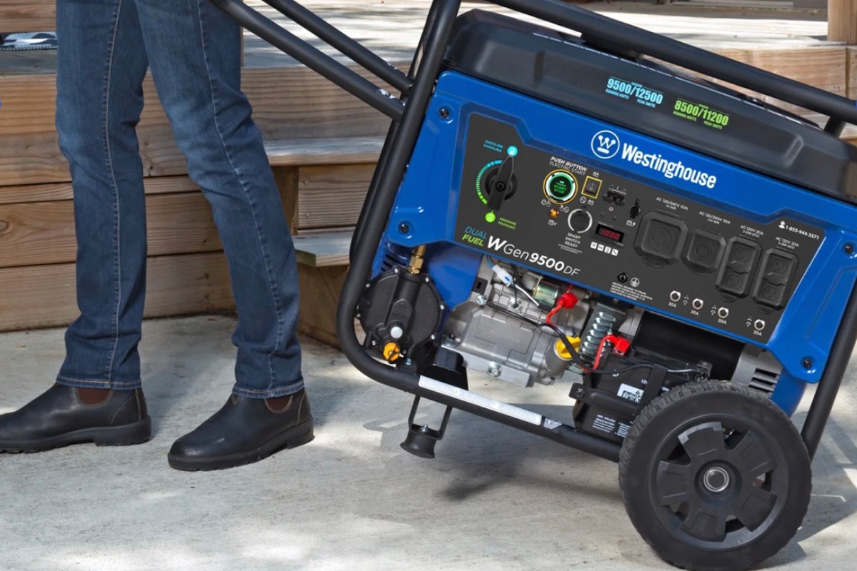 A person in jeans and black boots is maneuvering a blue Westinghouse dual fuel generator.