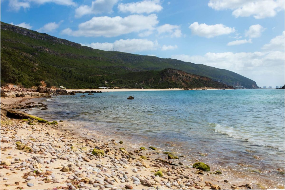A sandy beach with pebbles and rocky shoreline, surrounded by green hills under a blue sky with scattered clouds.