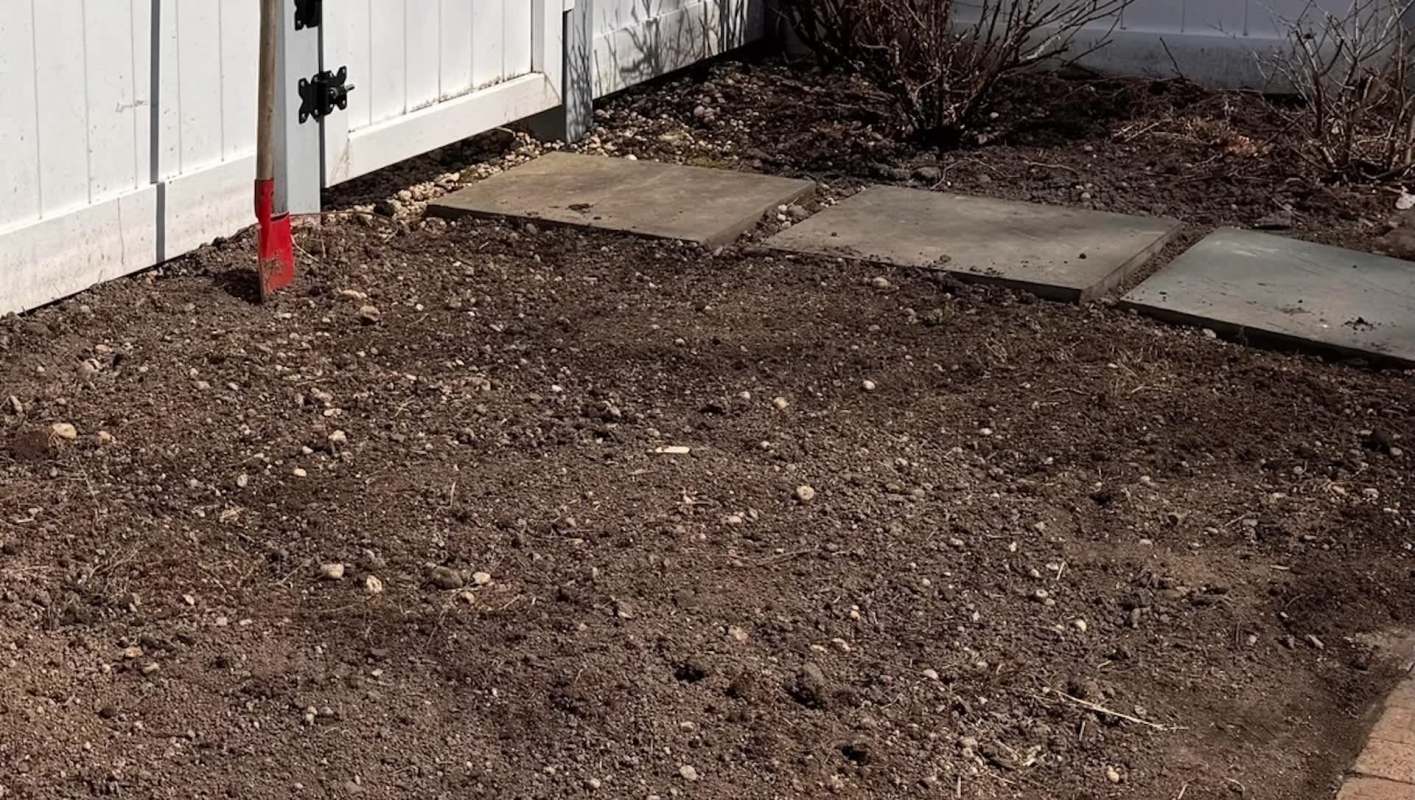 A garden area with freshly turned soil, a red-handled shovel, and stone stepping stones beside a white fence.