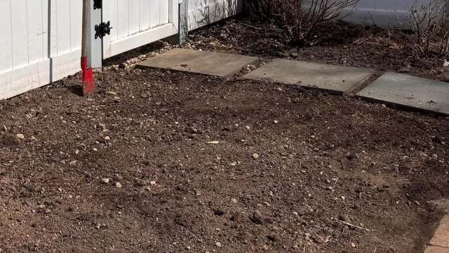 A garden area with freshly turned soil, a red-handled shovel, and stone stepping stones beside a white fence.