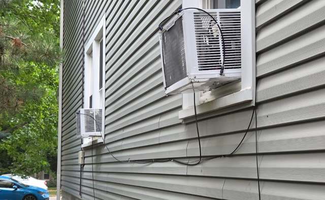 Two air conditioning units mounted on a gray siding wall, with electrical wires visible.