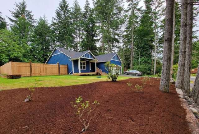 A blue house with a wooden fence surrounded by trees and freshly mulched garden beds.