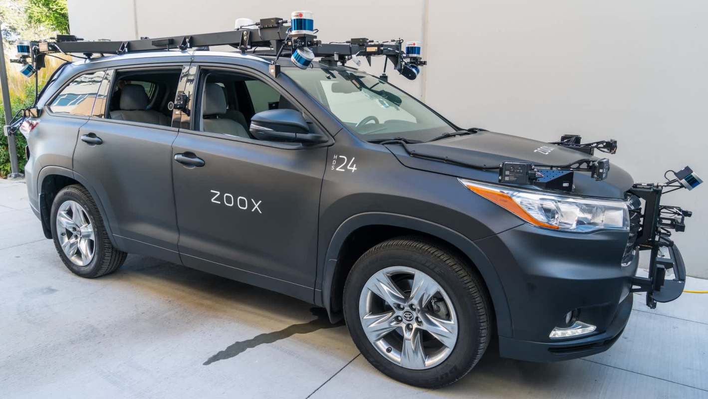 A black vehicle with "Zoox" on the side, equipped with sensors and cameras for autonomous driving research.