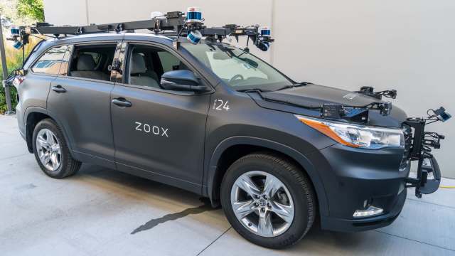A black vehicle with "Zoox" on the side, equipped with sensors and cameras for autonomous driving research.