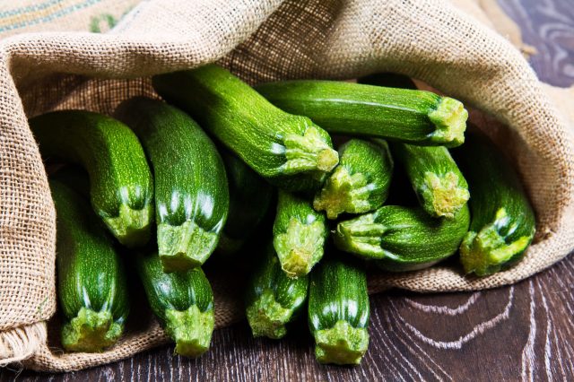 A burlap sack contains fresh, green zucchinis resting on a wooden surface.