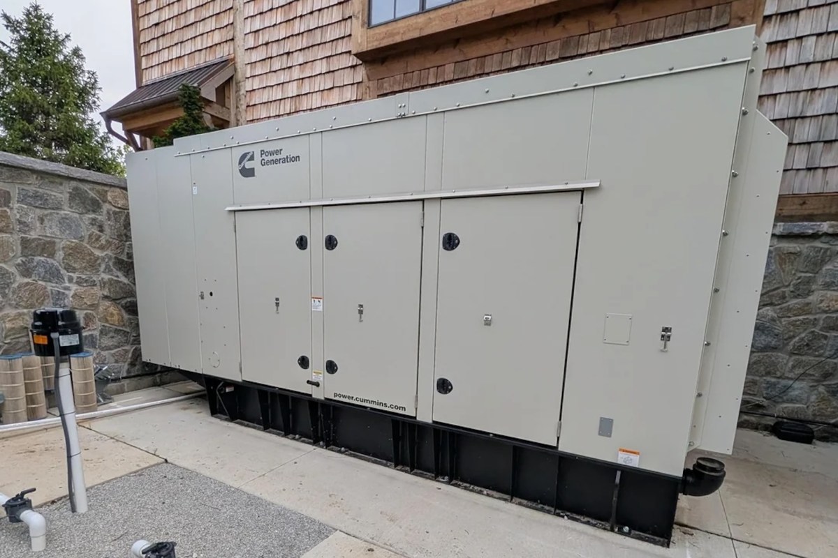 A large generator unit placed beside a stone wall and a house with wooden slding.