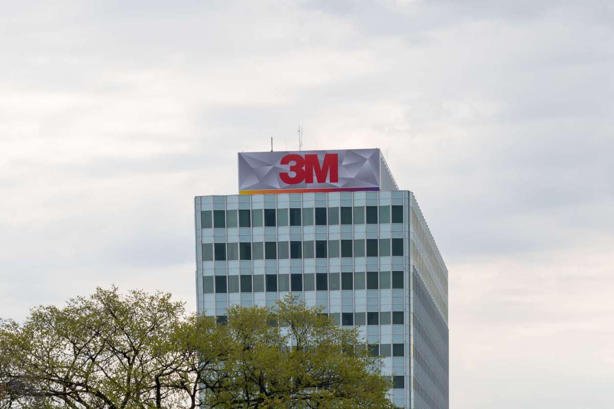 A building with the 3M logo on the rooftop, partially obscured by trees and under a cloudy sky.