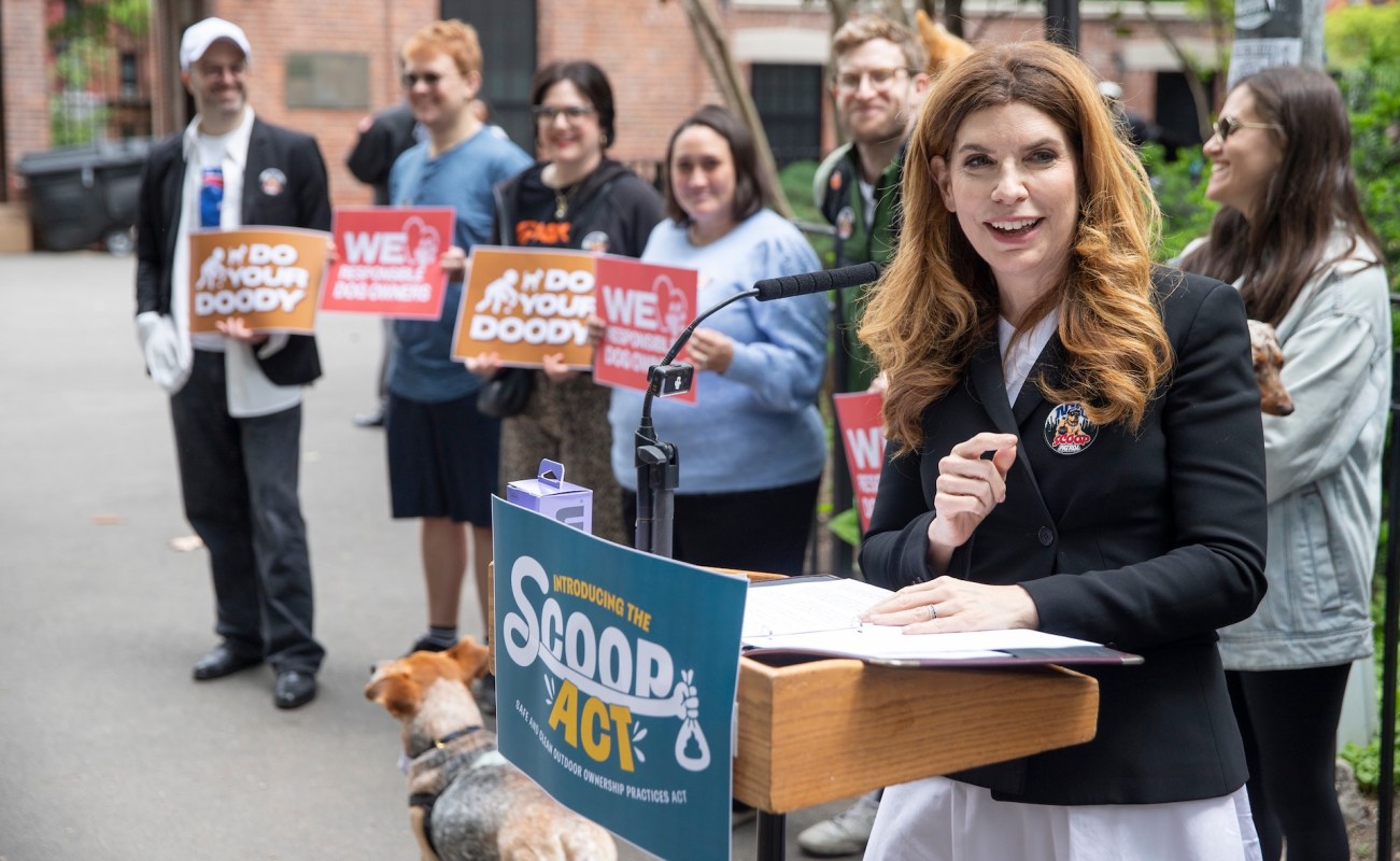 A woman speaks at a podium with supporters holding signs in the background advocating for responsible dog ownership.