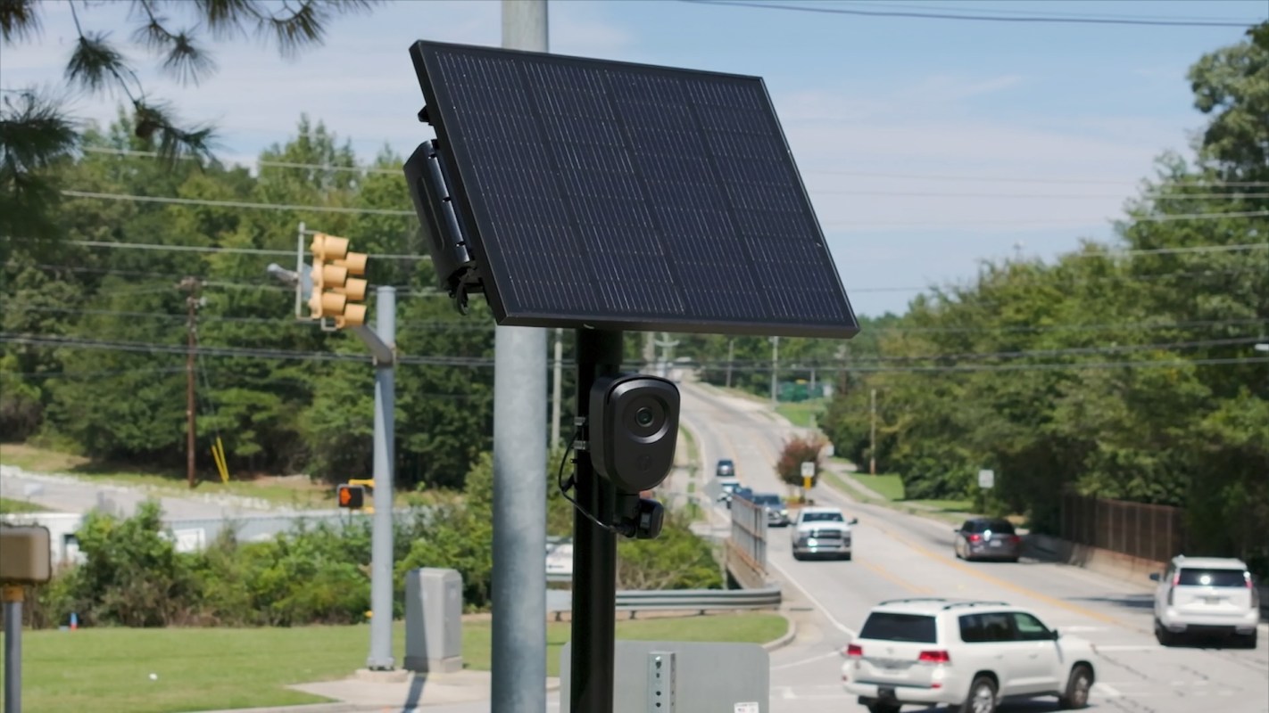 A Flock camera on a pole beside a road with vehicles passing by.