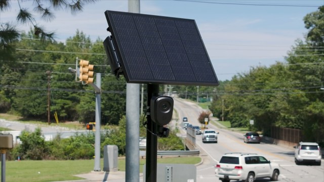 A Flock camera on a pole beside a road with vehicles passing by.