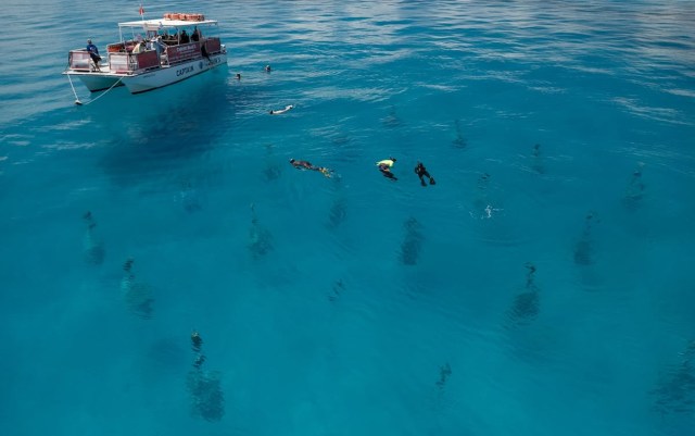 A boat floats on clear blue water with several people snorkeling nearby around vertical columns of coral reef.