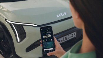 A person holds a smartphone displaying an app for a Kia vehicle parked nearby.