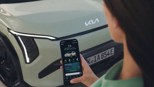 A person holds a smartphone displaying an app for a Kia vehicle parked nearby.