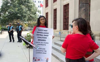 A group protests with signs demanding the cancellation of surveillance contracts with Flock outside a government building.