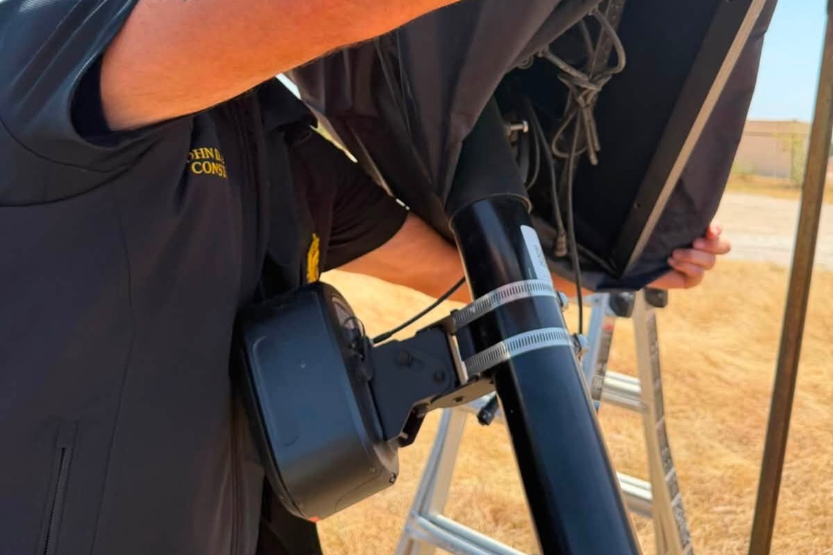 A person covers a Flock Safety camera on a pole with a covering while standing on a ladder in a dry outdoor environment.