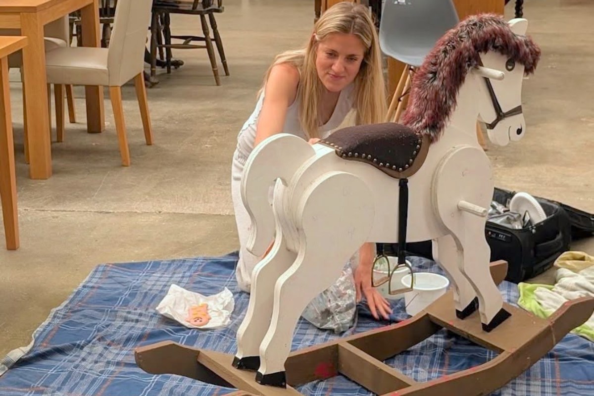 A woman adjusts a rocking horse on a blue plaid blanket in a large indoor space.