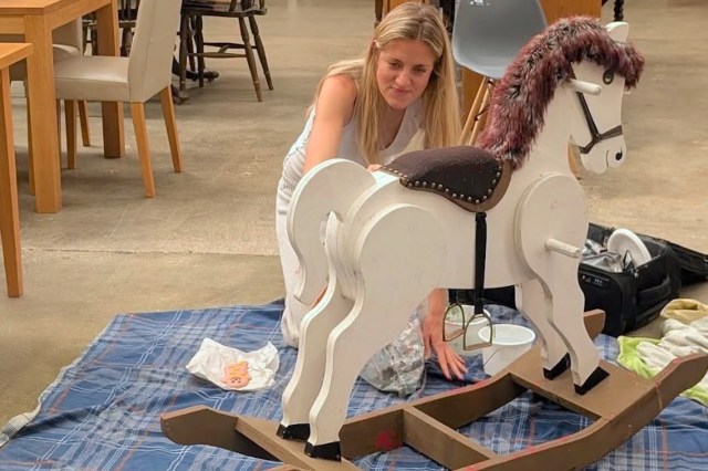 A woman adjusts a rocking horse on a blue plaid blanket in a large indoor space.
